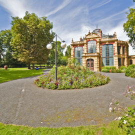 Das Bild zeigt einen gepflegten Garten mit einem runden Weg, der ein zentrales Blumenbeet umgibt. Im Hintergrund steht ein prunkvolles Gebäude mit großen Rundbogenfenstern und kunstvollen architektonischen Details. Umgeben ist die Szene von grünen Bäumen und einem leicht bewölkten Himmel.