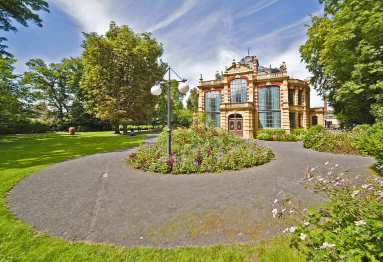 Das Bild zeigt einen gepflegten Garten mit einem runden Weg, der ein zentrales Blumenbeet umgibt. Im Hintergrund steht ein prunkvolles Gebäude mit großen Rundbogenfenstern und kunstvollen architektonischen Details. Umgeben ist die Szene von grünen Bäumen und einem leicht bewölkten Himmel.