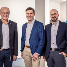 Das Bild zeigt drei Personen in Businesskleidung, die nebeneinander stehen. Links steht eine Person im dunklen Anzug mit hellem Hemd, in der Mitte eine Person mit blauem Sakko, weißem Hemd und beiger Hose, rechts eine weitere Person im dunklen Anzug mit hellem Hemd. Im Hintergrund befindet sich ein Banner mit Informationen zu Bau- und Prüfdienstleistungen sowie eine Pflanze auf der linken Seite.