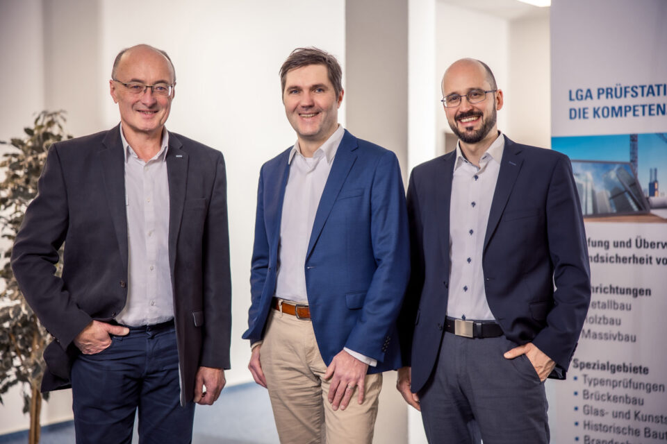 Das Bild zeigt drei Personen in Businesskleidung, die nebeneinander stehen. Links steht eine Person im dunklen Anzug mit hellem Hemd, in der Mitte eine Person mit blauem Sakko, weißem Hemd und beiger Hose, rechts eine weitere Person im dunklen Anzug mit hellem Hemd. Im Hintergrund befindet sich ein Banner mit Informationen zu Bau- und Prüfdienstleistungen sowie eine Pflanze auf der linken Seite.