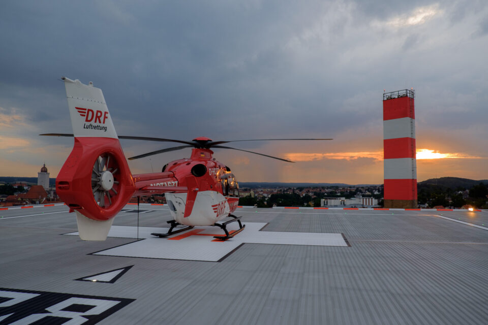 Das Bild zeigt einen rot-weißen Rettungshubschrauber auf einem Landeplatz. Im Hintergrund ist eine Stadtlandschaft bei Sonnenuntergang zu sehen, mit Wolken am Himmel und einem hohen rot-weiß gestreiften Turm.