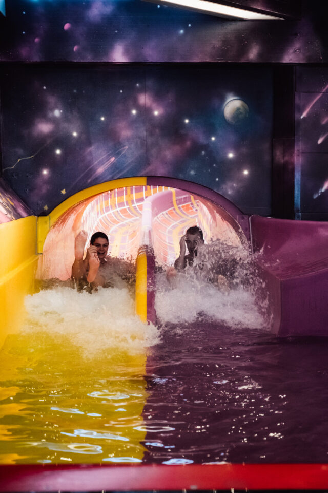 Das Bild zeigt zwei Personen, die nebeneinander eine Wasserrutsche hinunter in ein Becken rutschen. Die Rutsche ist zweigeteilt mit einer gelben und einer lilafarbenen Bahn. Im Hintergrund ist ein Wandbild mit Weltraummotiven wie Sternen und Planeten zu sehen.