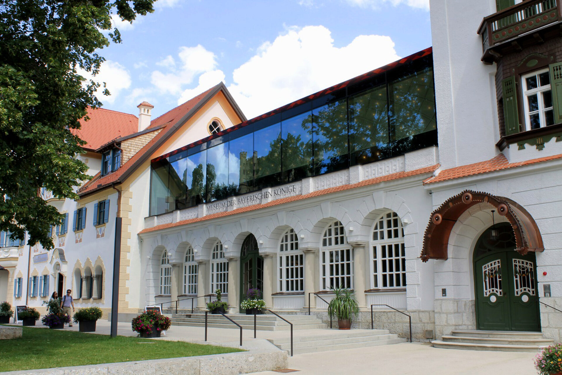 Das Bild zeigt ein Gebäude mit einer Kombination aus traditioneller und moderner Architektur. Der untere Teil hat eine weiße Fassade mit Rundbogenfenstern, während der obere Bereich durch eine moderne Glasaufstockung ergänzt wird. Vor dem Eingang stehen Blumentöpfe, und das Gebäude ist von Bäumen umgeben.