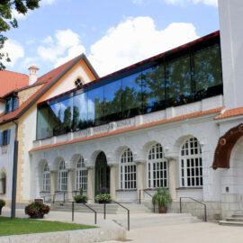 Das Bild zeigt ein Gebäude mit einer Kombination aus traditioneller und moderner Architektur. Der untere Teil hat eine weiße Fassade mit Rundbogenfenstern, während der obere Bereich durch eine moderne Glasaufstockung ergänzt wird. Vor dem Eingang stehen Blumentöpfe, und das Gebäude ist von Bäumen umgeben.