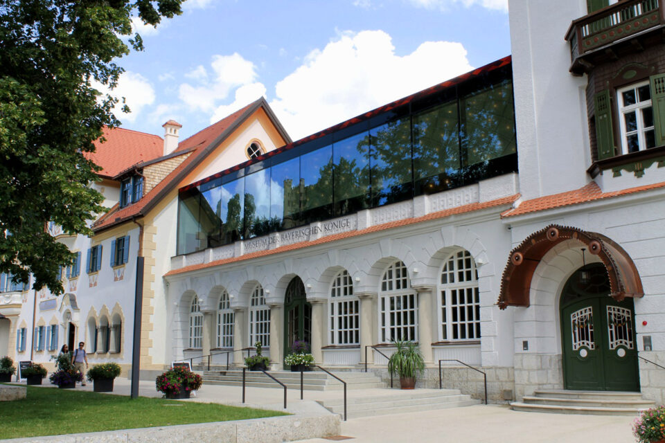 Das Bild zeigt ein Gebäude mit einer Kombination aus traditioneller und moderner Architektur. Der untere Teil hat eine weiße Fassade mit Rundbogenfenstern, während der obere Bereich durch eine moderne Glasaufstockung ergänzt wird. Vor dem Eingang stehen Blumentöpfe, und das Gebäude ist von Bäumen umgeben.