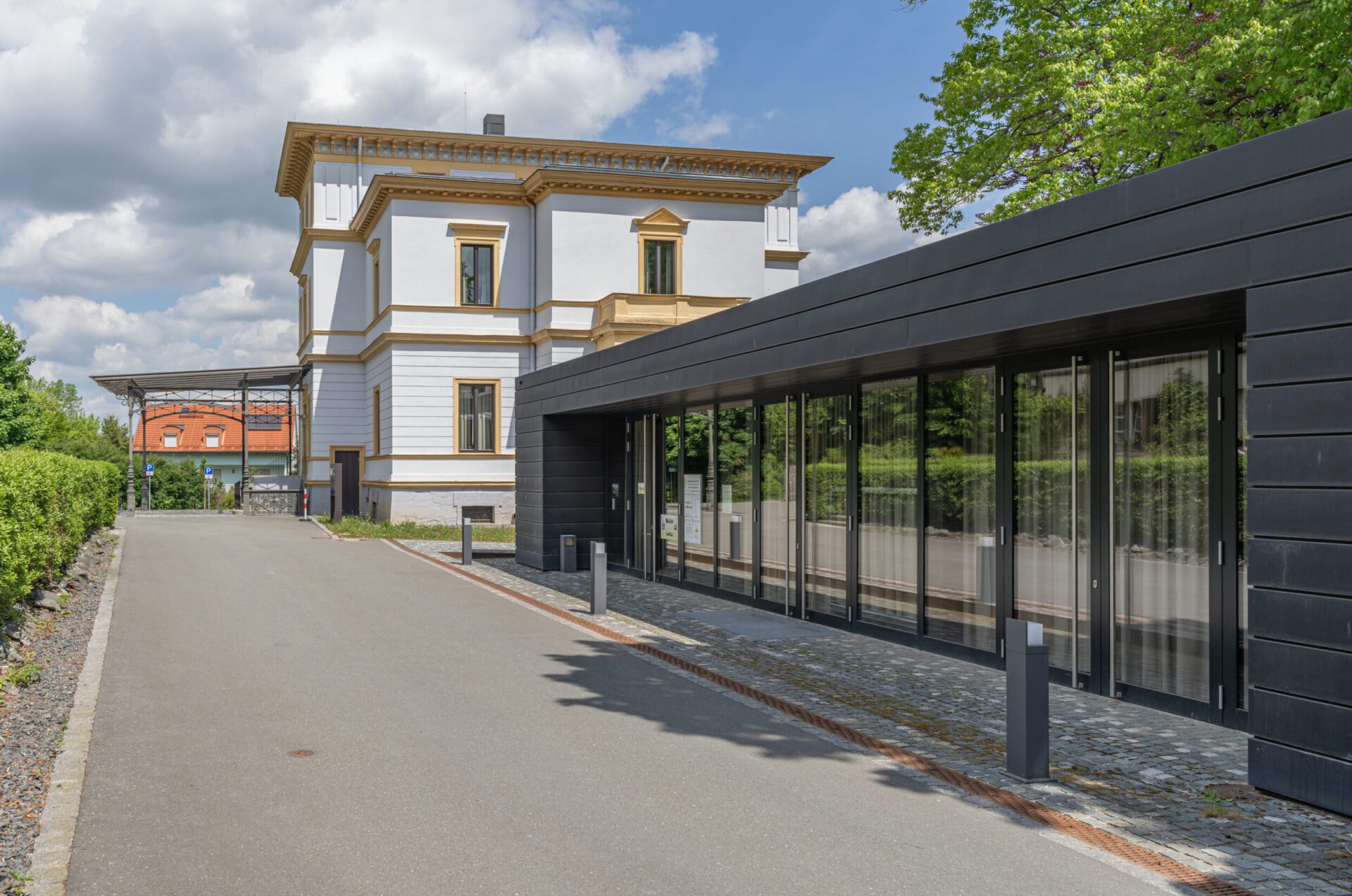 Das Bild zeigt rechts ein modernes Gebäude mit Glasfassade und links ein traditionelles Gebäude mit weißen Wänden und goldenen Verzierungen. Beide Gebäude sind durch einen Weg verbunden, der von Grünpflanzen gesäumt ist. Im Hintergrund sind Bäume zu sehen, der Himmel ist teils bewölkt.