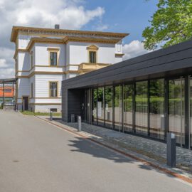 Das Bild zeigt rechts ein modernes Gebäude mit Glasfassade und links ein traditionelles Gebäude mit weißen Wänden und goldenen Verzierungen. Beide Gebäude sind durch einen Weg verbunden, der von Grünpflanzen gesäumt ist. Im Hintergrund sind Bäume zu sehen, der Himmel ist teils bewölkt.