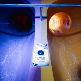 Das Bild zeigt zwei Wasserrutschen, eine in Blau und eine in Gelb beleuchtet. Über den Rutschen verläuft ein Metallgeländer. In der Mitte befindet sich ein großer Knopf.