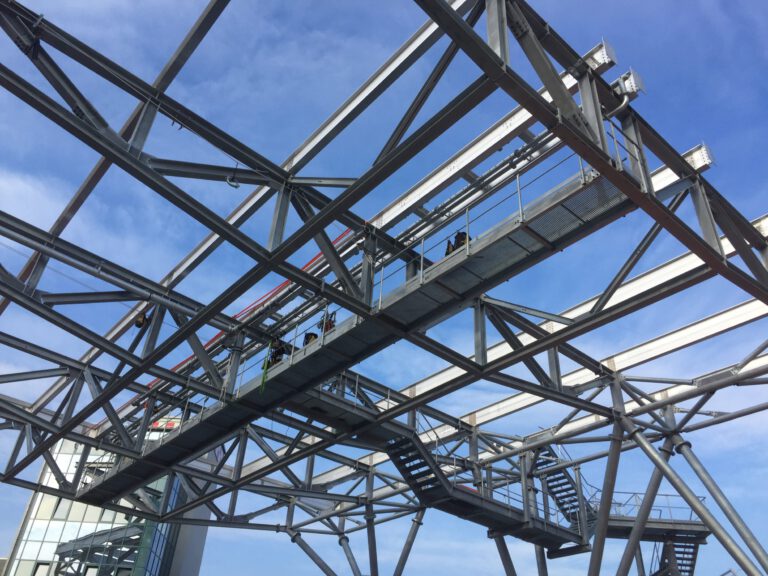 Das Bild zeigt eine komplexe Stahlkonstruktion mit zahlreichen sich kreuzenden Trägern und Streben. Eine begehbare Plattform mit Geländern und Treppen ist in die Struktur integriert. Der Hintergrund zeigt einen klaren blauen Himmel, was auf eine Außenaufnahme hinweist.