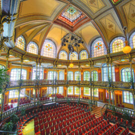 Das Bild zeigt den prunkvollen Innenraum eines historischen Theaters oder Konzertsaals mit hoher, kunstvoll verzierter Decke und bunten Glasfenstern. Rote Polstersitze sind halbkreisförmig im Parkett und auf einem verzierten Balkon angeordnet. Große Kronleuchter und dekorative Säulen unterstreichen die elegante Atmosphäre, ergänzt durch einige Topfpflanzen.