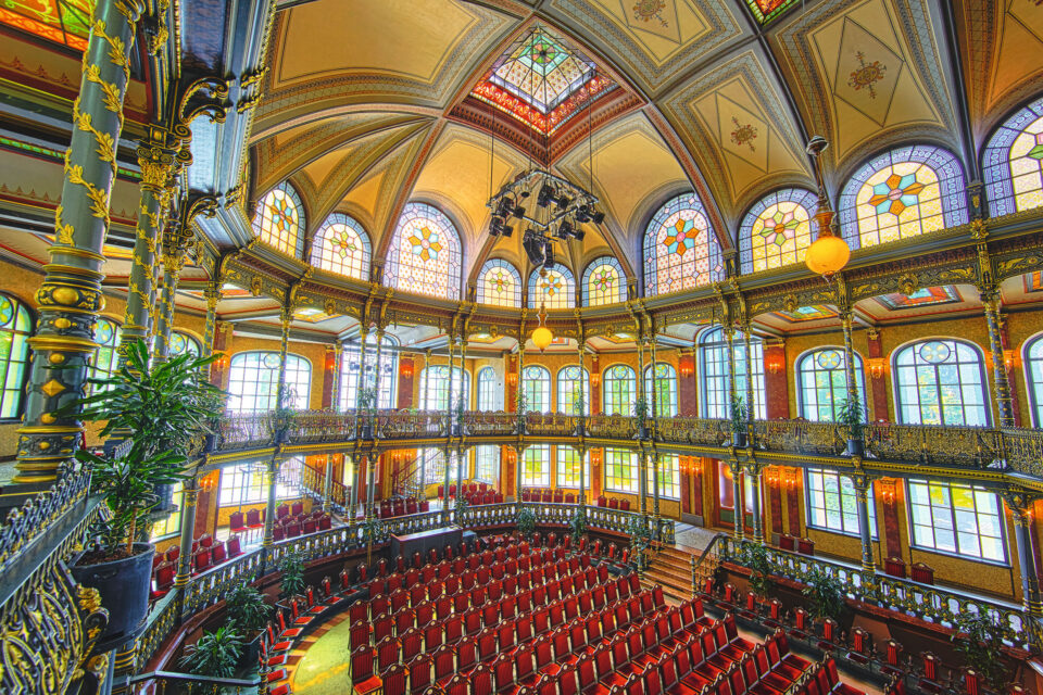 Das Bild zeigt den prunkvollen Innenraum eines historischen Theaters oder Konzertsaals mit hoher, kunstvoll verzierter Decke und bunten Glasfenstern. Rote Polstersitze sind halbkreisförmig im Parkett und auf einem verzierten Balkon angeordnet. Große Kronleuchter und dekorative Säulen unterstreichen die elegante Atmosphäre, ergänzt durch einige Topfpflanzen.