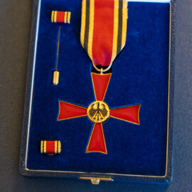 Das Bild zeigt das Bundesverdienstkreuz in seiner Präsentationsschatulle. Die Auszeichnung besteht aus einem rot emaillierten Kreuz mit goldenen Rändern und einem goldenen Medaillon mit dem Bundesadler in der Mitte. In der Schatulle befinden sich außerdem ein Miniaturband und eine Anstecknadel.