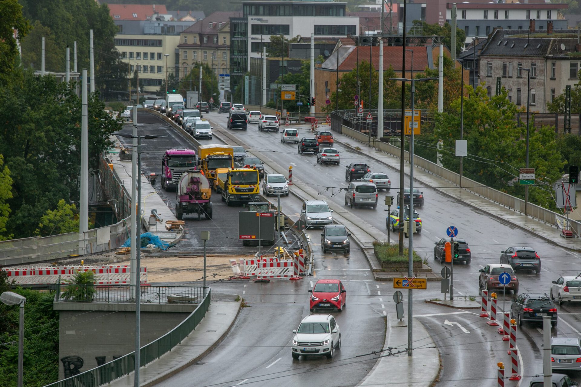 Das Bild zeigt eine stark befahrene städtische Straße mit mehreren Fahrspuren, auf denen Autos und Lastwagen unterwegs sind. Auf der linken Seite findet eine Straßenbaumaßnahme statt, erkennbar an Baustellenfahrzeugen und Absperrungen. Der nasse Straßenbelag und Lichtreflexionen deuten auf regnerisches Wetter hin, im Hintergrund sind Gebäude und Bäume sichtbar.