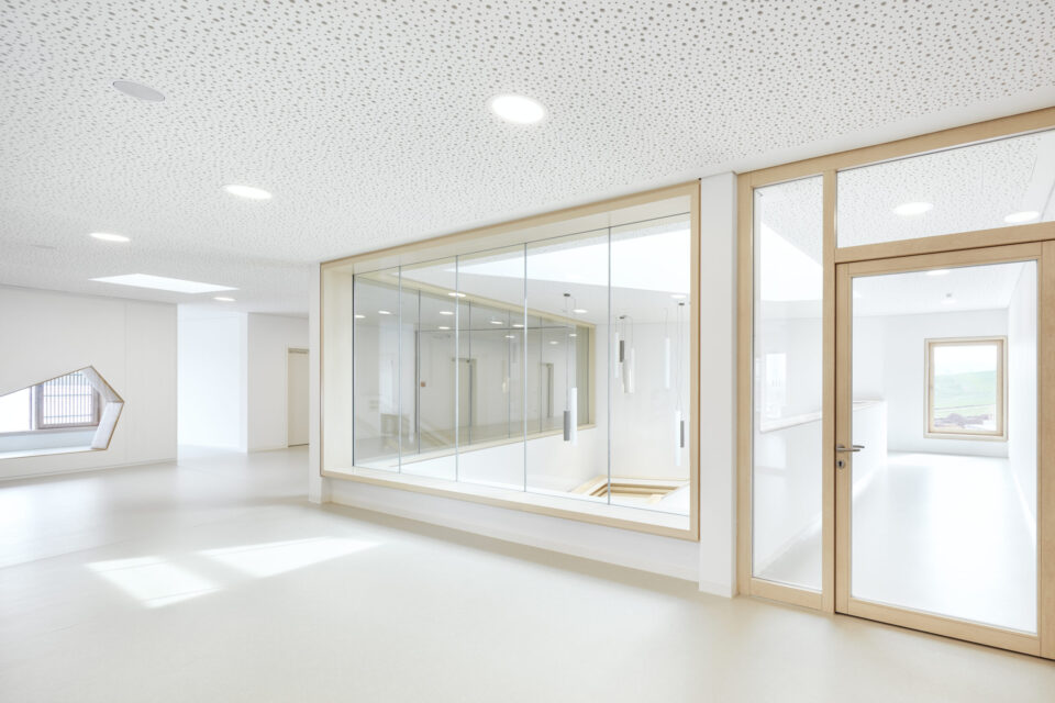 Das Bild zeigt einen modernen, minimalistisch gestalteten Innenraum mit weißen Wänden und einer Decke mit Punktmuster und eingelassener Beleuchtung. In der Mitte befindet sich ein großes Glasfenster mit Blick in einen angrenzenden Raum, rechts daneben eine Tür mit Holzrahmen und Glaseinsatz. Links ist eine in die Wand integrierte, eckige Fenstersitzbank zu sehen.