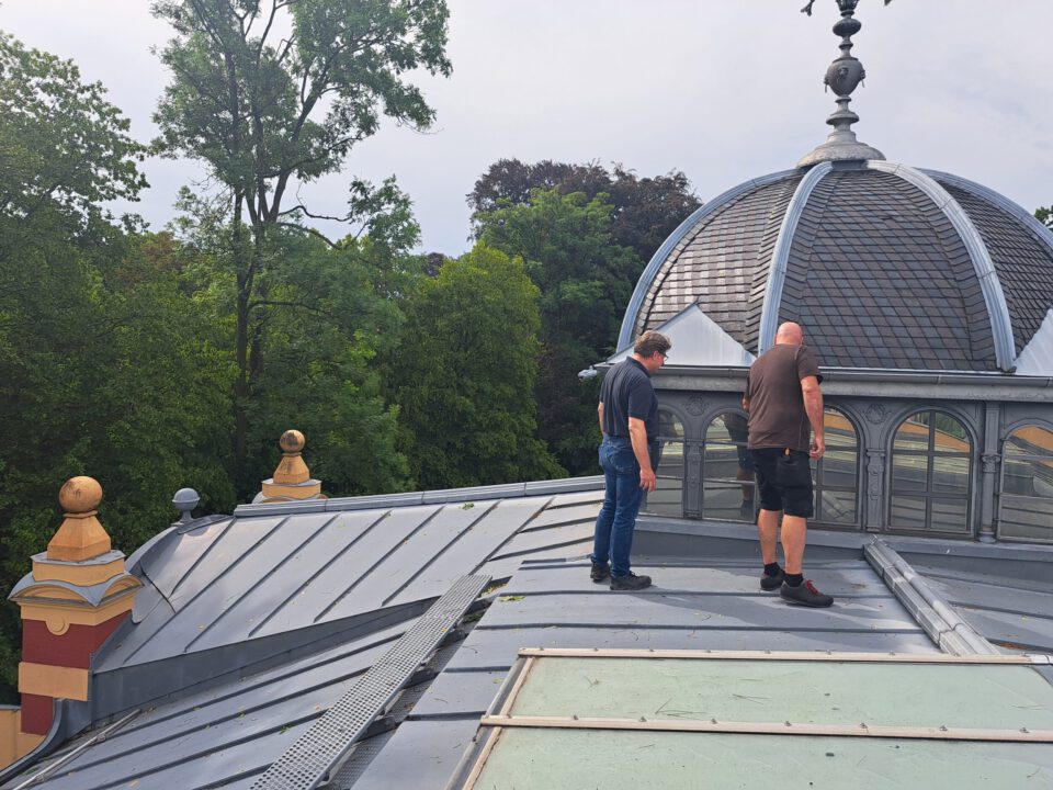 Das Bild zeigt zwei Personen auf dem Dach eines Gebäudes mit einer kuppelförmigen Metallstruktur und Fenstern. Die Personen scheinen das Dach zu inspizieren oder daran zu arbeiten. Im Hintergrund sind Bäume zu sehen, die auf eine grüne Umgebung hinweisen.