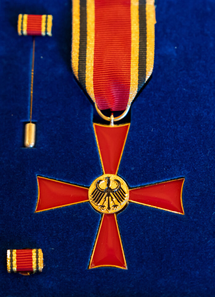Das Bild zeigt das Bundesverdienstkreuz in seiner Präsentationsschatulle. Die Auszeichnung besteht aus einem rot emaillierten Kreuz mit goldenen Rändern und einem goldenen Medaillon mit dem Bundesadler in der Mitte. In der Schatulle befinden sich außerdem ein Miniaturband und eine Anstecknadel.