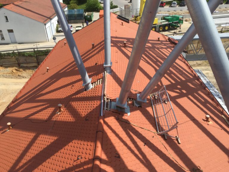 Das Bild zeigt ein rotes Ziegeldach, durch das mehrere große Metallträger verlaufen. Die Träger werfen Schatten auf das Dach und erzeugen ein auffälliges Muster. Im Hintergrund sind weitere Gebäude, Baumaschinen und Fahrzeuge zu sehen.