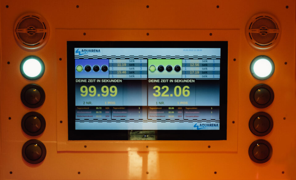 Das Bild zeigt einen Bildschirm mit mehreren Zahlenanzeigen und einem orangefarbenen Rahmen mit Knöpfen und Lautsprechern. Auf dem Display sind zwei Zeitmessungen in Sekunden sowie weitere kleinere Zeiten dargestellt. Der Bildschirm gehört zu einer Anlage zur Zeitmessung, vermutlich im Bereich Wasserrutschen.