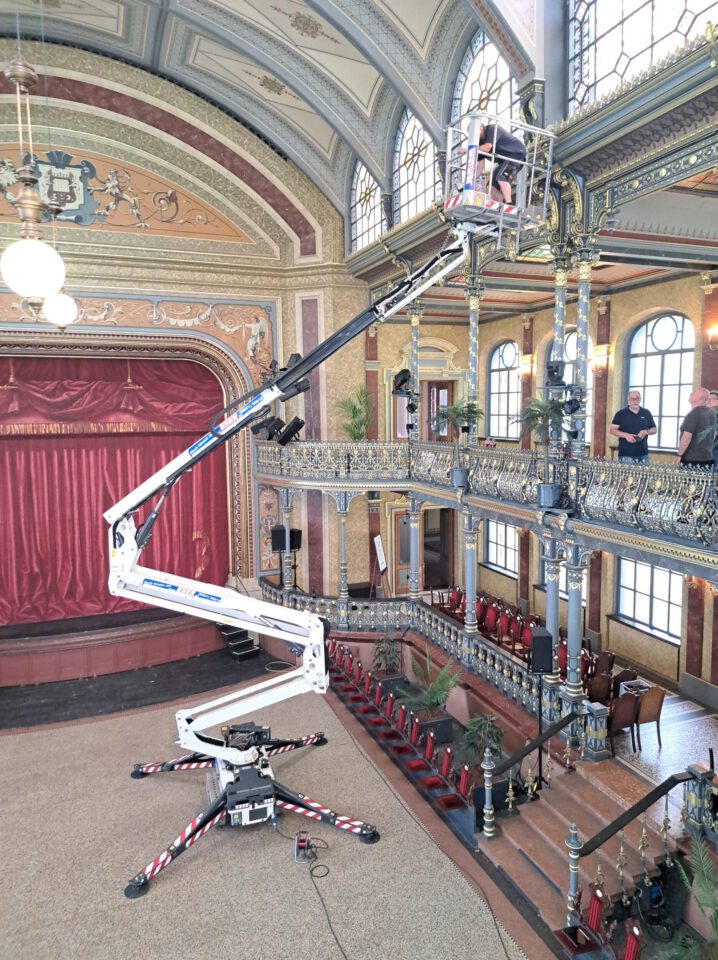 Das Bild zeigt einen historischen Innenraum mit hoher Decke, großen Fenstern und kunstvollen architektonischen Details wie Säulen und Verzierungen. Links befindet sich eine Bühne mit rotem Vorhang, davor steht ein mechanischer Lift, auf dem eine Person in der oberen Etage arbeitet. Eine weitere Person beobachtet die Szene vom zweiten Stock aus.