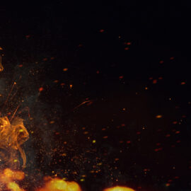 Das Bild zeigt eine Nahaufnahme von Flammen vor einem dunklen Hintergrund. Die Feuerzungen sind leuchtend gelb und orange, mit Funken und Glutpartikeln, die in der Luft schweben.