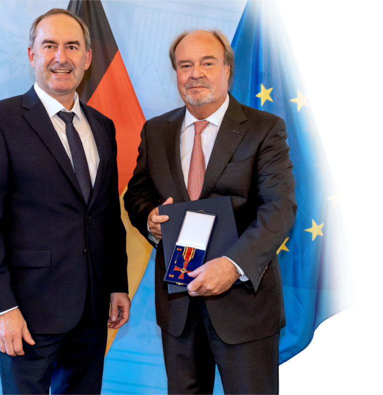 Zwei Männer im Anzug stehen nebeneinander. Der Mann rechts hält eine geöffnete Schachtel mit einer Medaille oder Auszeichnung. Im Hintergrund sind die Flaggen von Deutschland und der Europäischen Union zu sehen.