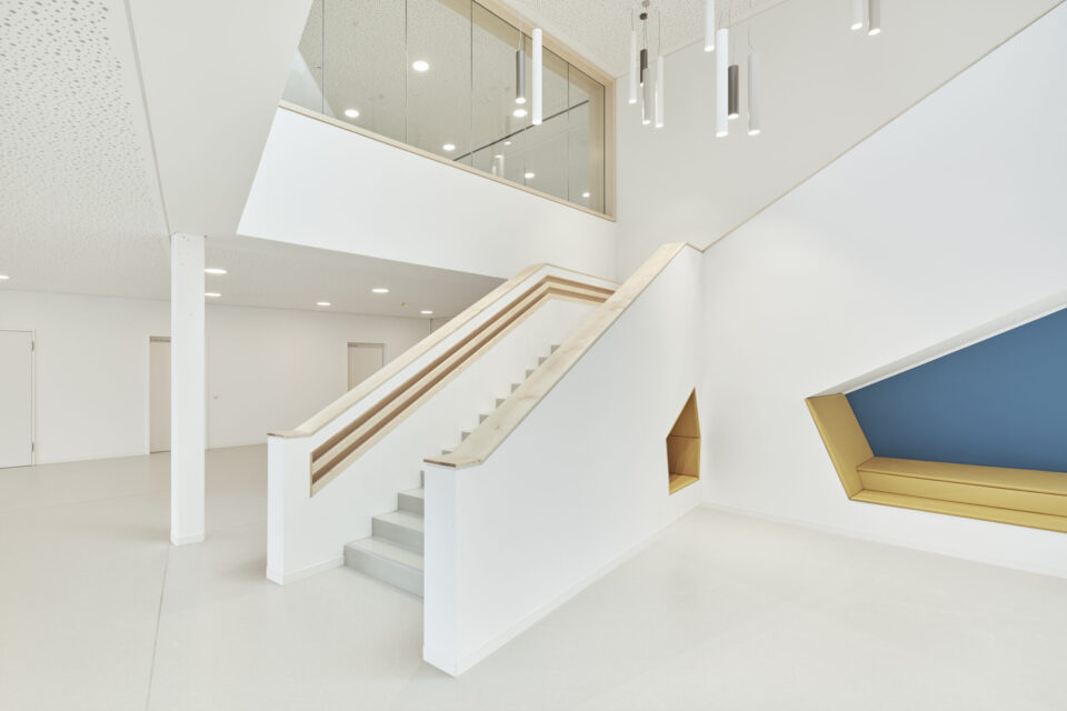 Das Bild zeigt einen modernen, hellen Innenraum mit weißen Wänden und Decken sowie einer Treppe mit Holzhandlauf, die zu einer oberen Ebene mit Glasbrüstung führt. An der Decke befinden sich eingelassene Leuchten und zylindrische Hängelampen. Rechts ist eine in die Wand integrierte Sitznische mit gelben Polstern und blauer Rückenlehne zu sehen.