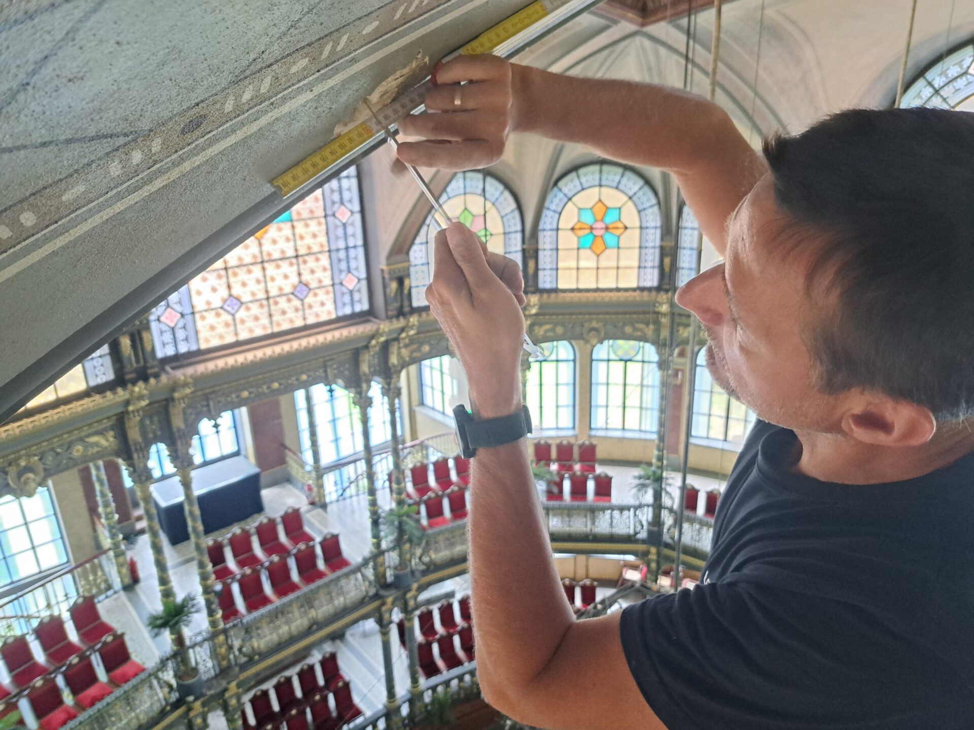 Das Bild zeigt eine Person, die mit einem gelben Maßband und einem Werkzeug etwas an der Decke eines prunkvollen Gebäudes misst. Im Hintergrund sind mehrere Ebenen mit roten Stühlen sowie große Buntglasfenster zu sehen.