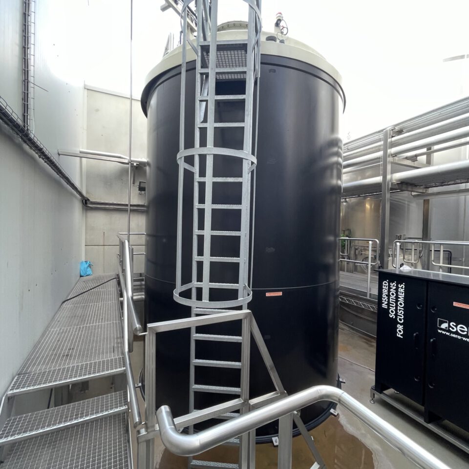 Das Bild zeigt eine industrielle Anlage mit einem großen, schwarzen, zylindrischen Tank, an dem eine Leiter befestigt ist. Um den Tank herum befinden sich Metallstege mit Geländern sowie verschiedene Rohre und technische Geräte im Hintergrund.