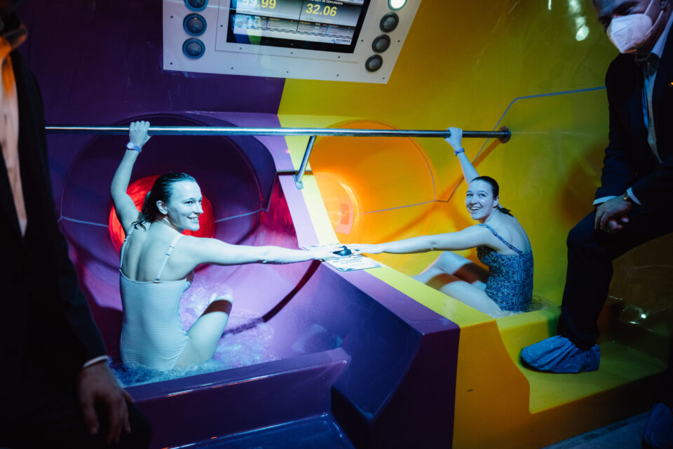 Das Bild zeigt zwei Personen, die am Einstieg zweier Wasserrutschen sitzen, eine in Lila und eine in Gelb. Sie reichen sich über eine Trennwand hinweg die Hände. Ein Mann steht daneben und beobachtet die Szene, über den Rutschen ist eine Anzeigetafel mit Zahlen zu sehen.