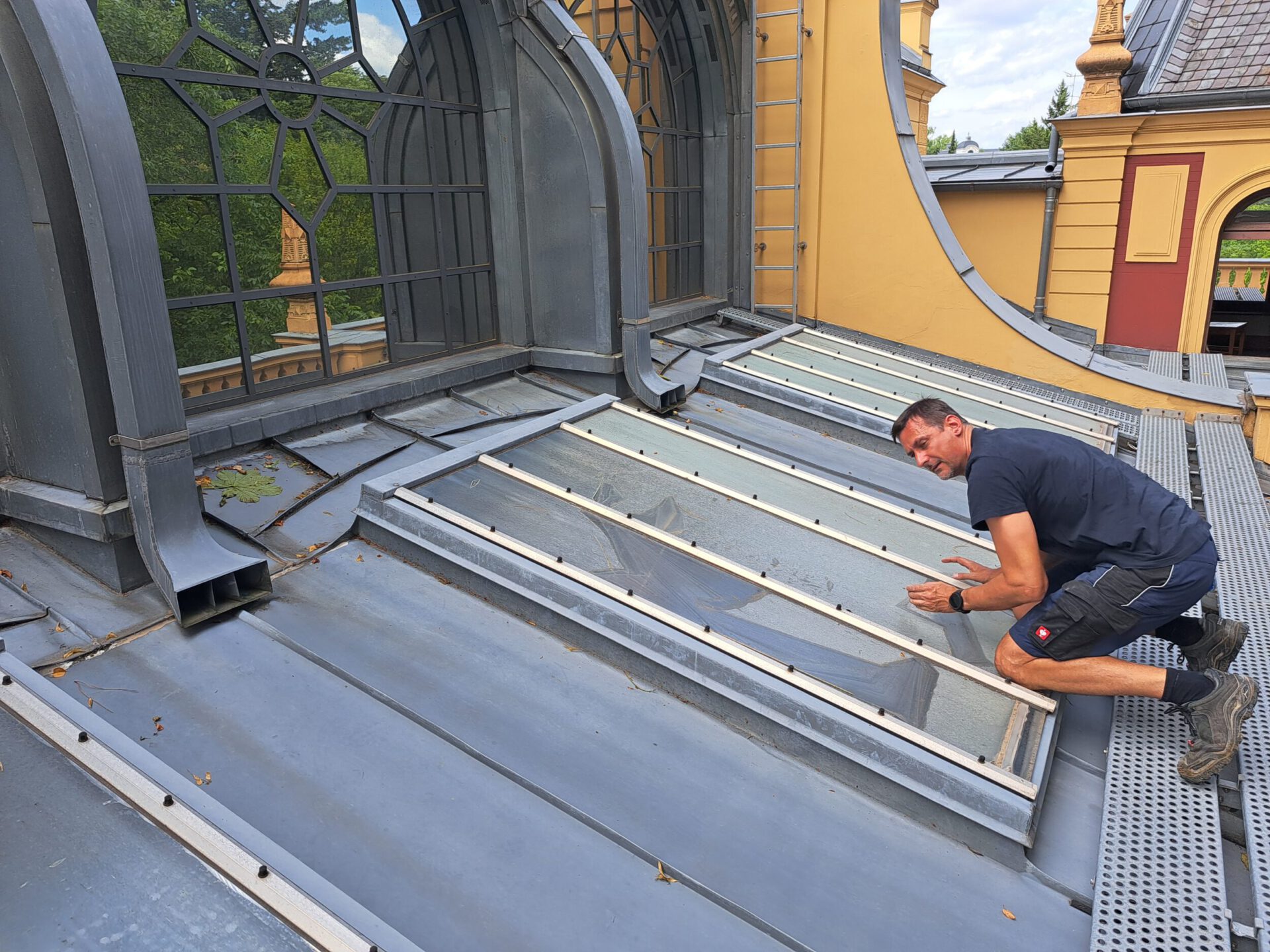 Das Bild zeigt eine Person, die auf einem Metalldach kniet und an den darin eingebetteten Glasflächen arbeitet oder diese inspiziert. Das Gebäude hat große Rundbogenfenster mit dekorativen Metallrahmen und ist gelb gestrichen. Durch die Fenster sind Bäume zu sehen, was auf eine grüne Umgebung hinweist.