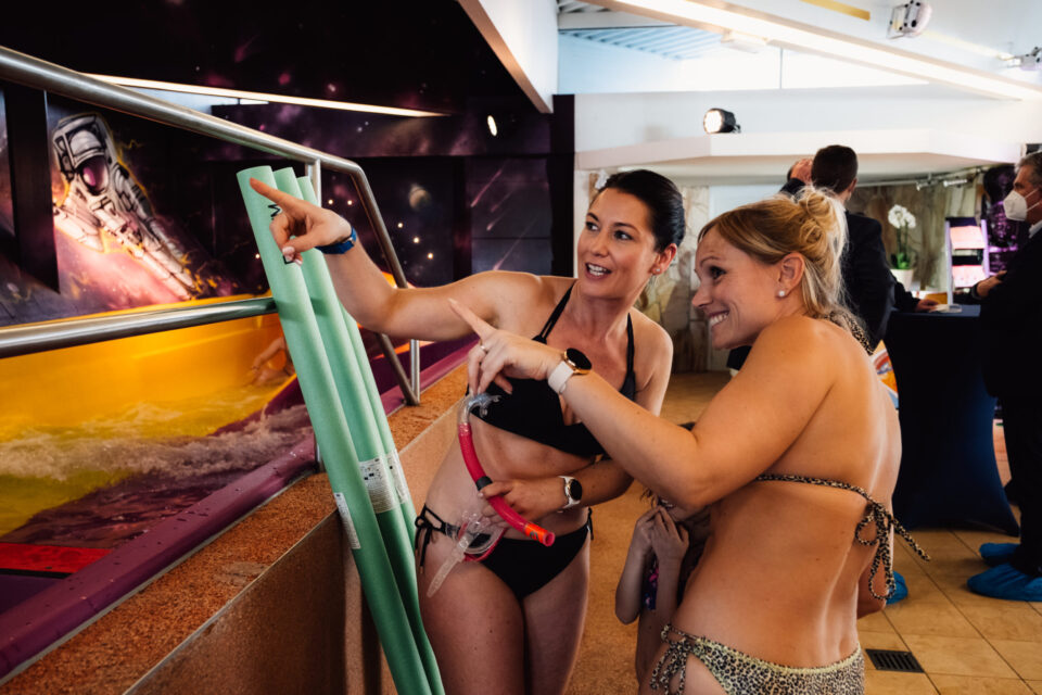 Zwei Personen in Badekleidung deuten nach links. Neben ihnen an ein Becken angelehnt 3 Schwimmnudeln. Im Hintergrund weitere Personen.