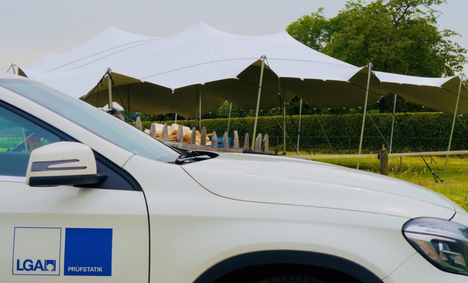 Das Bild zeigt ein weißes Fahrzeug, das vor einer großen Zeltkonstruktion im Freien geparkt ist. Das Zelt wird von mehreren Masten getragen und steht auf einer Wiese mit Bäumen und Hecken im Hintergrund. Unter dem Zelt befinden sich verschiedene Gegenstände, möglicherweise Baumaterialien oder Geräte.