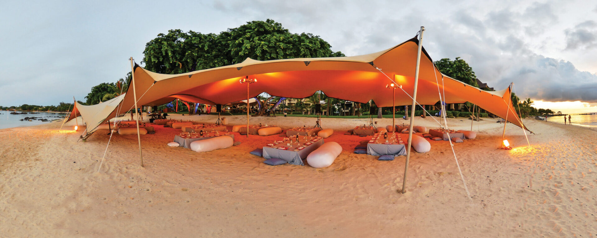 Das Bild zeigt eine Strandszene mit einem großen, beigefarbenen Baldachinzelt, das auf dem Sand aufgestellt ist. Unter dem Zelt stehen niedrige Tische mit Tischdecken, umgeben von Sitzkissen, und die Tische sind mit Tellern, Gläsern und Kerzen gedeckt. Im Hintergrund sind Bäume und etwas Grün zu sehen, links im Bild das Meer, darüber ein wolkiger Himmel mit Lichtstimmung von Sonnenauf- oder -untergang.