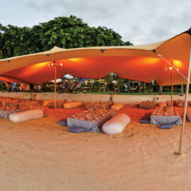 Das Bild zeigt eine Strandszene mit einem großen, beigefarbenen Baldachinzelt, das auf dem Sand aufgestellt ist. Unter dem Zelt stehen niedrige Tische mit Tischdecken, umgeben von Sitzkissen, und die Tische sind mit Tellern, Gläsern und Kerzen gedeckt. Im Hintergrund sind Bäume und etwas Grün zu sehen, links im Bild das Meer, darüber ein wolkiger Himmel mit Lichtstimmung von Sonnenauf- oder -untergang.