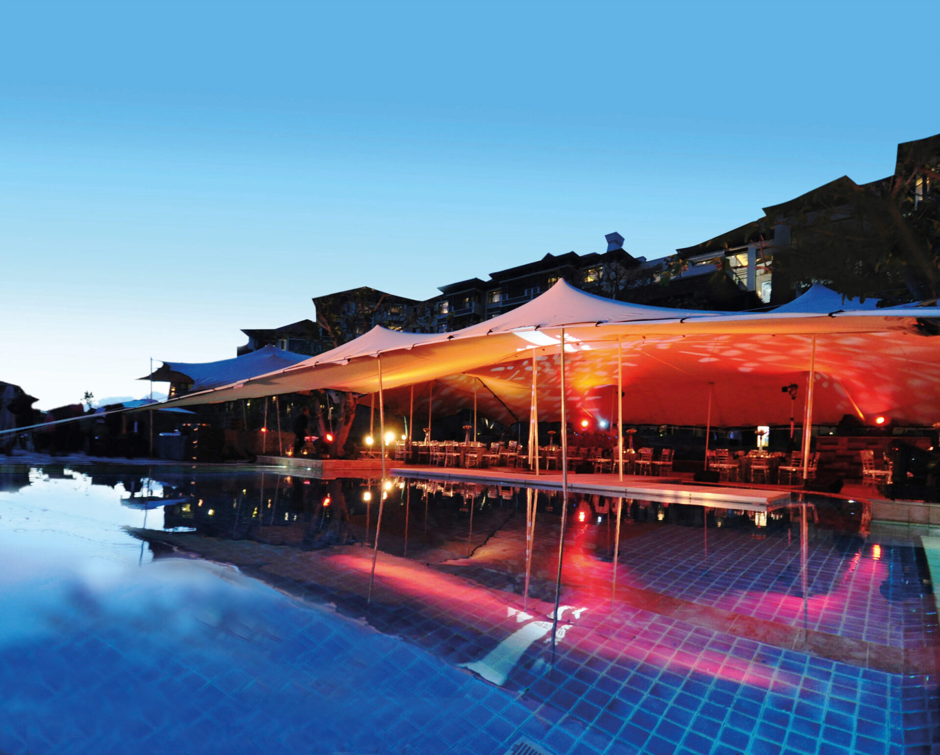 Das Bild zeigt eine große Zeltkonstruktion im Freien, die mit farbigen Lichtern in Rot und Gelb beleuchtet ist. Das Zelt steht neben einem Swimmingpool mit klarem, blauem Wasser, das die Lichter und die Struktur reflektiert. Im Hintergrund sind Gebäude im Abendlicht zu sehen, unter dem Zelt stehen Tische und Stühle für eine Veranstaltung.