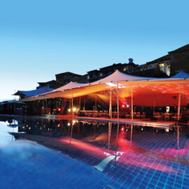 Das Bild zeigt eine große Zeltkonstruktion im Freien, die mit farbigen Lichtern in Rot und Gelb beleuchtet ist. Das Zelt steht neben einem Swimmingpool mit klarem, blauem Wasser, das die Lichter und die Struktur reflektiert. Im Hintergrund sind Gebäude im Abendlicht zu sehen, unter dem Zelt stehen Tische und Stühle für eine Veranstaltung.
