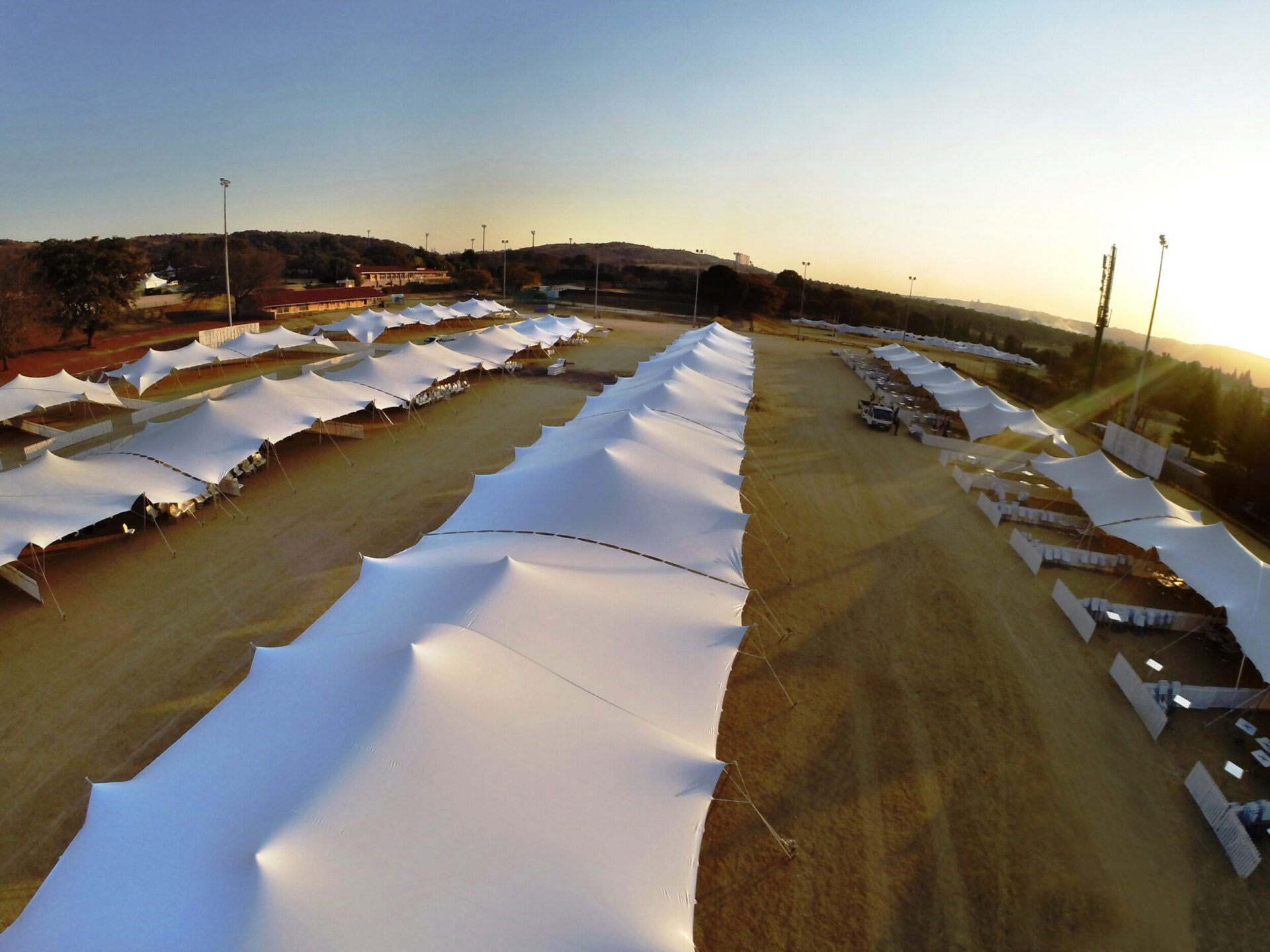 Das Bild zeigt ein großes Außengelände mit mehreren Reihen weißer Zelte, die auf sandigem oder erdigem Boden aufgestellt sind. Die Zelte sind in parallelen Linien angeordnet und bilden ein symmetrisches Muster. Die Aufnahme stammt aus erhöhter Perspektive bei tiefstehender Sonne, wodurch lange Schatten entstehen, im Hintergrund sind Bäume, Gebäude und Lichtmasten zu sehen.