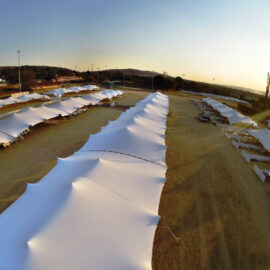 Das Bild zeigt ein großes Außengelände mit mehreren Reihen weißer Zelte, die auf sandigem oder erdigem Boden aufgestellt sind. Die Zelte sind in parallelen Linien angeordnet und bilden ein symmetrisches Muster. Die Aufnahme stammt aus erhöhter Perspektive bei tiefstehender Sonne, wodurch lange Schatten entstehen, im Hintergrund sind Bäume, Gebäude und Lichtmasten zu sehen.