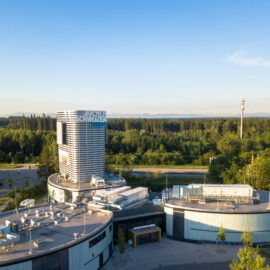 Das Bild zeigt ein modernes Gebäudekomplex inmitten von Grünflächen und Bäumen. Im Zentrum steht ein hoher, zylindrischer Turm mit einem Schriftzug am oberen Rand. Umgeben ist der Turm von flachen Gebäuden mit technischen Anlagen auf den Dächern sowie Straßen und Wegen, im Hintergrund erstreckt sich ein dichter Wald bis zum Horizont.