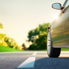 Das Bild zeigt eine Nahaufnahme eines Autos, das auf einer Straße fährt. Der Fokus liegt auf dem rechten Vorderrad, das sich in Bewegung befindet. Die Straße hat eine weiße gestrichelte Mittellinie, und an den Seiten sind grüne Bäume und Büsche zu sehen. Der Hintergrund ist leicht verschwommen, was auf Bewegung oder Geschwindigkeit hinweist.