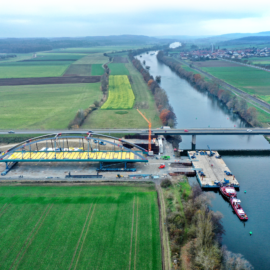 Das Bild zeigt eine Luftaufnahme einer Baustelle, auf der eine Brücke über einen Fluss errichtet wird. Die Brücke befindet sich noch im frühen Bauzustand, und große Kräne sowie Baumaschinen sind zu sehen. Umgeben ist die Baustelle von grünen Feldern, einem parallel verlaufenden Gewässer und weiter entfernten Gebäuden.