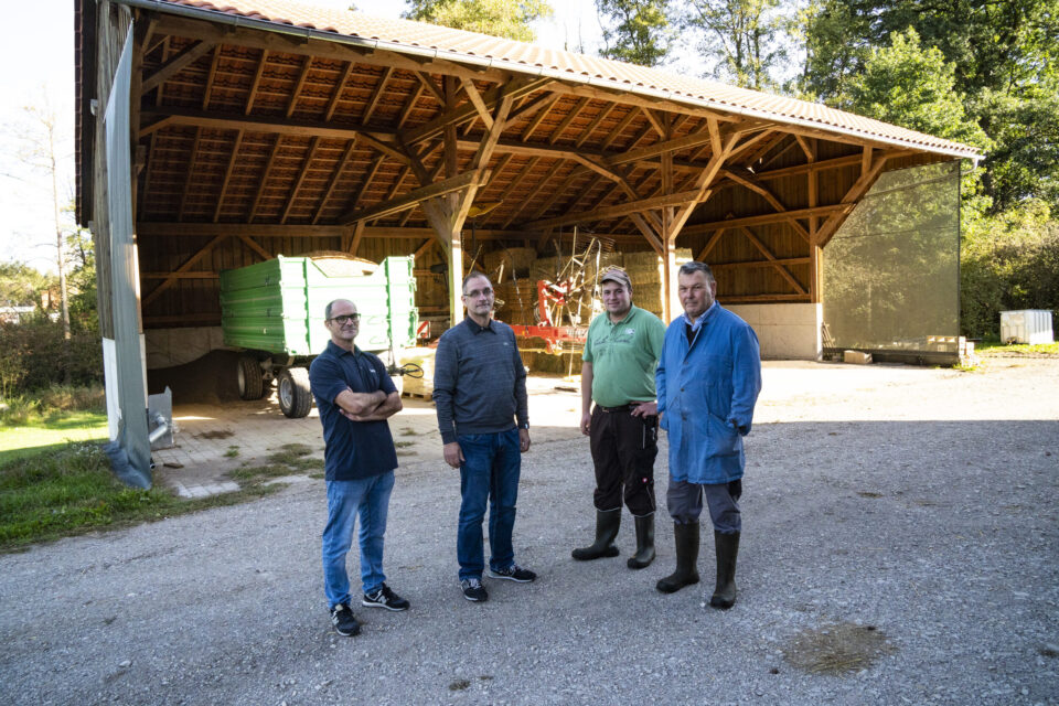 Das Bild zeigt vier Personen, die vor einer großen, offenen Scheune mit Holzrahmen und Ziegeldach stehen. In der Scheune befinden sich landwirtschaftliche Geräte, darunter ein grüner Anhänger. Eine der Personen trägt einen blauen Mantel und Gummistiefel, was auf eine Tätigkeit in der Landwirtschaft hinweist.