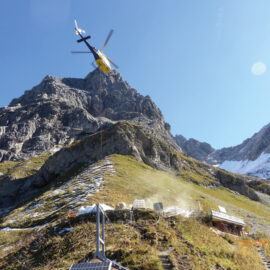 Das Bild zeigt eine alpine Landschaft mit einem gelb-schwarzen Hubschrauber, der über einem Berghang schwebt. Der Hubschrauber transportiert Ausrüstung oder Material an einem Seil. Auf dem Boden sind eine Wiese mit Schneeflecken sowie verschiedene Geräte oder Strukturen zu sehen.