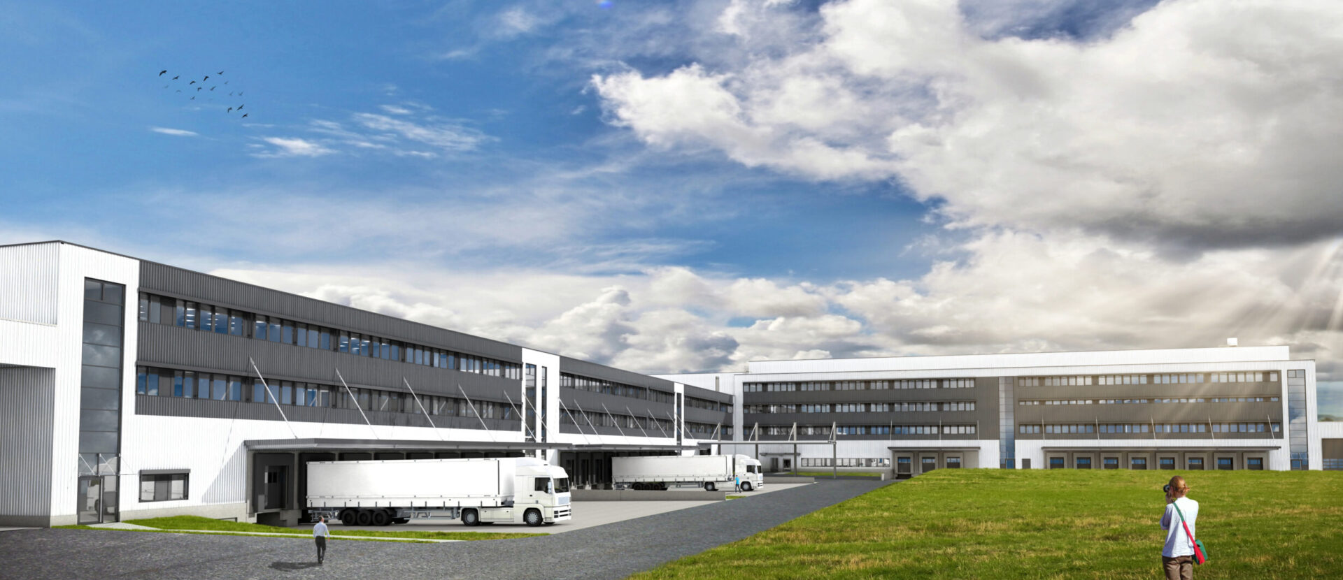 Das Bild zeigt einen großen Industrie- oder Gewerbebau mit mehreren Laderampen, an denen weiße Lastwagen geparkt sind. Das Gebäude hat mehrere Etagen mit vielen Fenstern. Im Vordergrund befindet sich eine Grasfläche, auf der zwei Personen spazieren gehen.