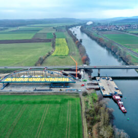 Das Bild zeigt eine Luftaufnahme einer Brückenbaustelle über einem Fluss. Auf der linken Seite befindet sich eine große Bogenkonstruktion, und auf der Baustelle sind Kräne, Fahrzeuge und Arbeiter zu sehen. Im Hintergrund verlaufen Felder, Straßen und eine weitere Brücke über den Fluss.
