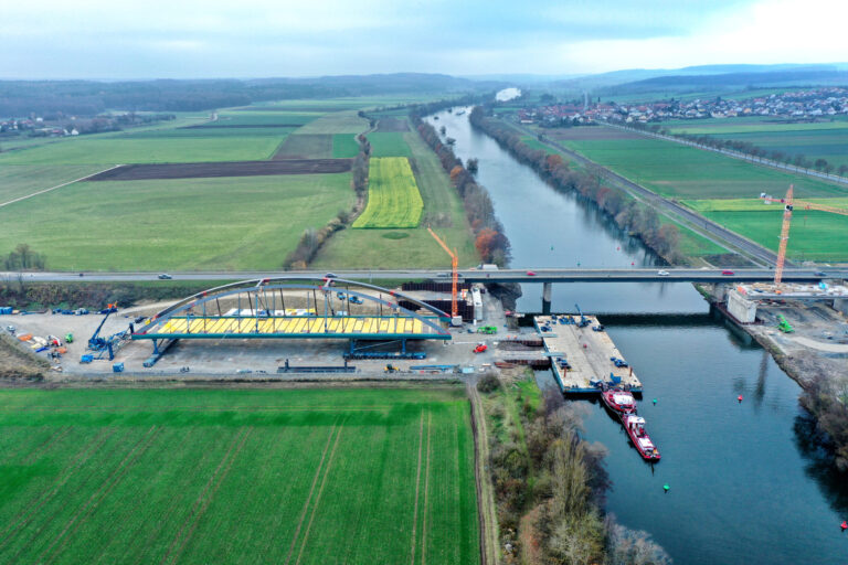 Das Bild zeigt eine Luftaufnahme einer Brückenbaustelle über einem Fluss. Auf der linken Seite befindet sich eine große Bogenkonstruktion, und auf der Baustelle sind Kräne, Fahrzeuge und Arbeiter zu sehen. Im Hintergrund verlaufen Felder, Straßen und eine weitere Brücke über den Fluss.