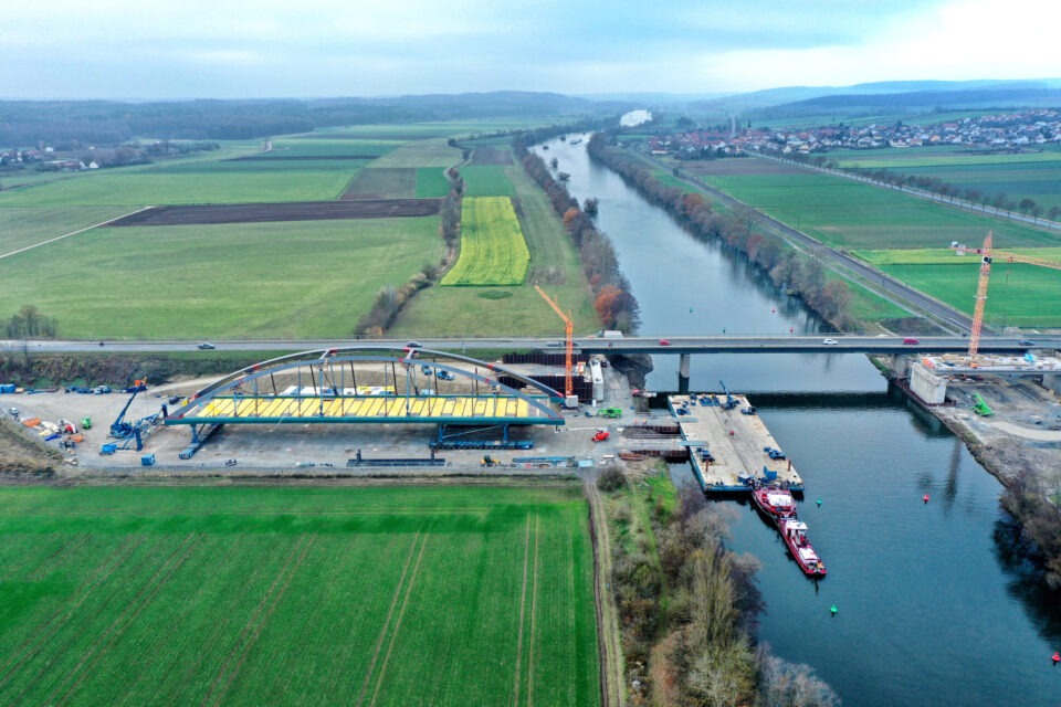 Das Bild zeigt eine Luftaufnahme einer Brückenbaustelle über einem Fluss. Auf der linken Seite befindet sich eine große Bogenkonstruktion, und auf der Baustelle sind Kräne, Fahrzeuge und Arbeiter zu sehen. Im Hintergrund verlaufen Felder, Straßen und eine weitere Brücke über den Fluss.
