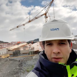 Das Bild zeigt eine Baustelle mit einem großen Kran im Hintergrund, der Baumaterialien hebt. Mehrere Arbeiter sind auf dem Gelände zu sehen, im Vordergrund steht eine Person mit weißem Schutzhelm und Warnjacke. Der Himmel ist teils bewölkt, im Hintergrund sind Hügel zu erkennen.