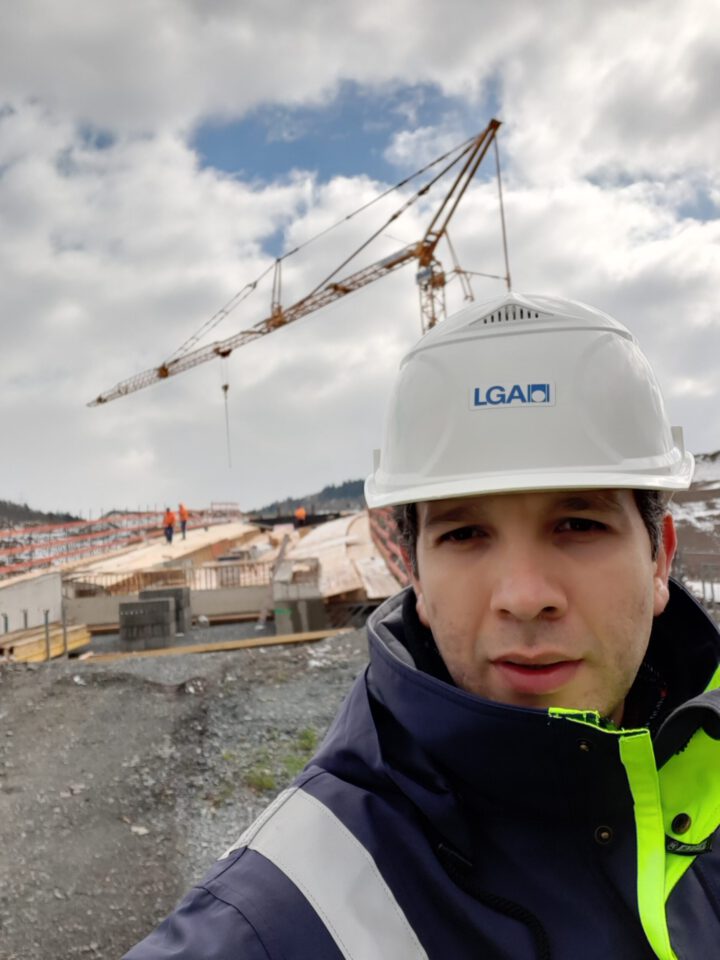 Das Bild zeigt eine Baustelle mit einem großen Kran im Hintergrund, der Baumaterialien hebt. Mehrere Arbeiter sind auf dem Gelände zu sehen, im Vordergrund steht eine Person mit weißem Schutzhelm und Warnjacke. Der Himmel ist teils bewölkt, im Hintergrund sind Hügel zu erkennen.