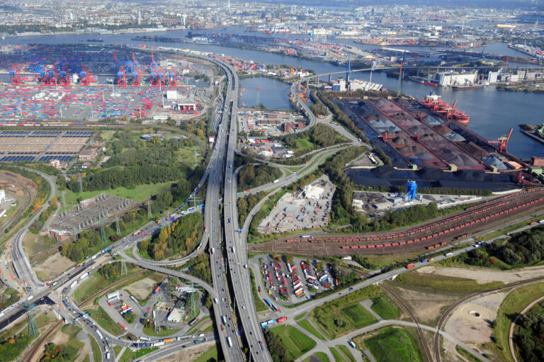 Das Bild zeigt eine Luftaufnahme eines großen Industriehafens mit zahlreichen Containern, Kränen und Frachtschiffen. Mehrere Straßen, Autobahnen und Gleise durchziehen das Gelände, das sich in der Nähe eines Gewässers befindet. Umgeben ist der Hafen von Industriegebäuden und vereinzelten Grünflächen.